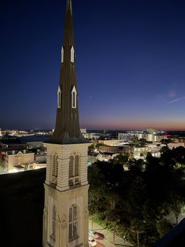 Aerial nighttime photograph of a tall church steeple silhouetted against a twilight sky with city lights visible in the background.
