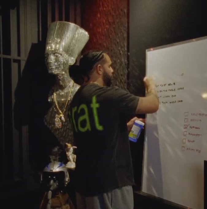 A man wearing a black t-shirt with green text writing on a whiteboard, standing next to a mirrored bust sculpture.
