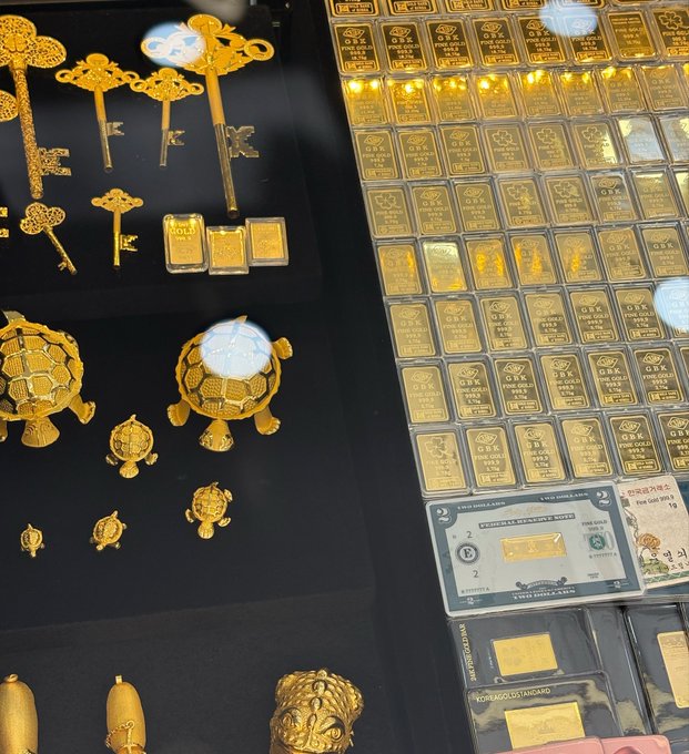 A display case showcasing gold items including decorative keys, small turtle figurines, and rows of gold bars labeled 'FINE GOLD 999.9' and '1 OZ GOLD BAR' with certificates.