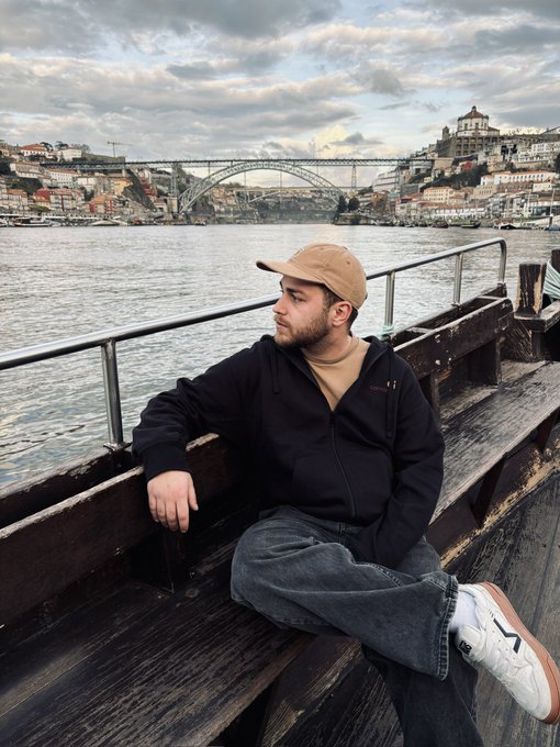 A young man in a black hoodie, beige cap, and white sneakers sitting on a wooden bench by a river, with a bridge and cityscape visible in the background.