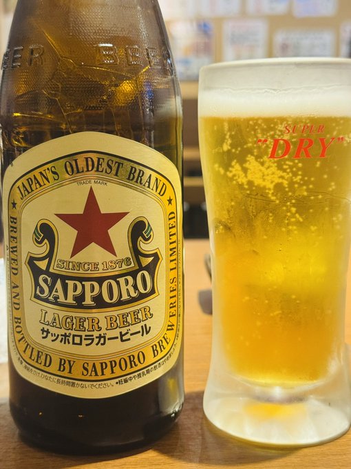 The image features a bottle of Sapporo beer next to a glass filled with beer, both placed on a table. The bottle has a label that says "Sapporo Lagrange Beer" written in Japanese. This suggests that the beer might be from Japan or is made by Sapporo Brewing Company. The glass of beer appears to have been poured and is ready for consumption.