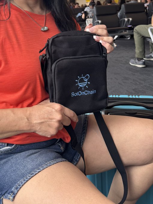 The image features a woman sitting down, holding a black shoulder bag that has a logo of "solonchain" printed on it. The bag appears to be made of canvas or nylon material with a black strap. In the background, there are other people present in what seems to be an airport setting.