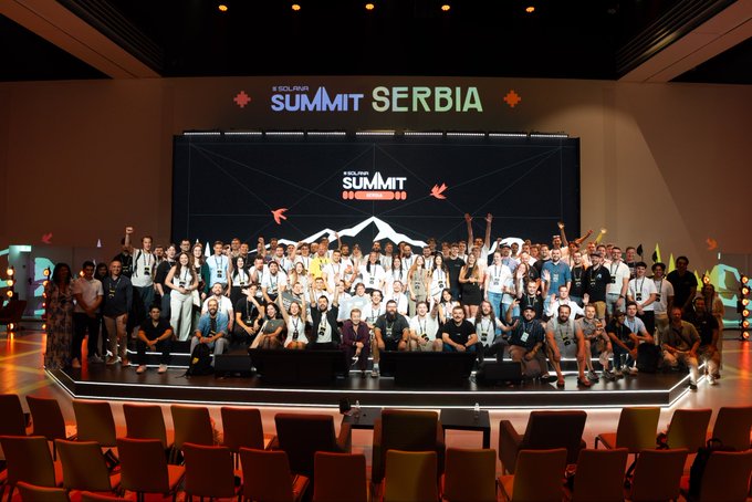 A large group photo of conference attendees posed on stage at the 'SOLANA SUMMIT SERBIA' event, with mountain graphics on the screen behind them.