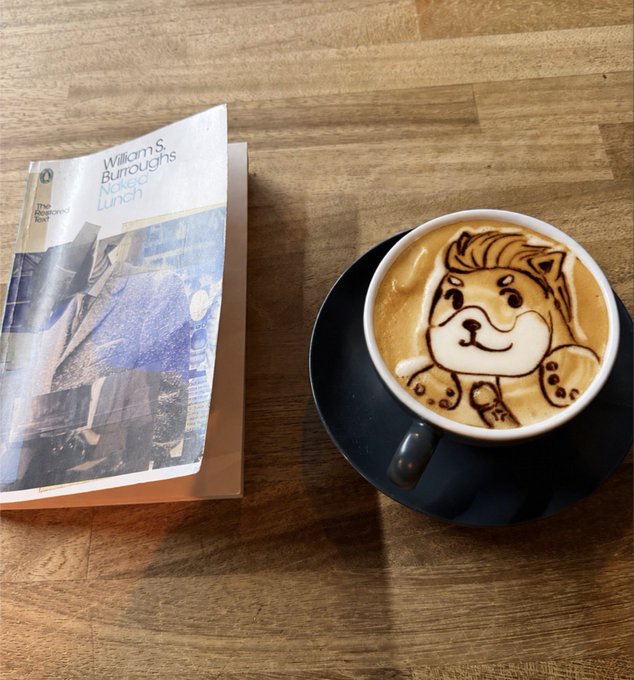 A latte art design featuring a cute dog face sits beside a paperback copy of William S. Burroughs' "Naked Lunch" on a wooden table, creating a cozy literary coffee break that pairs nicely with the playful "#CAW" caption. The whimsical puppy in the foam complements the relaxed reading vibe of the classic Beat novel.