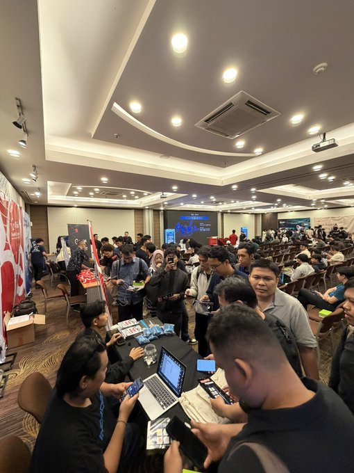 Crowded conference hall filled with attendees at the Indonesia Web3 Hidden Gems event, where people gather around booths using laptops and phones, with a projector screen visible in the background.