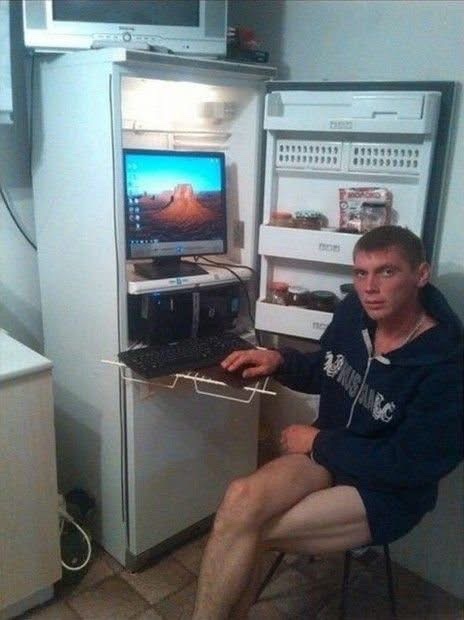 A photo of a man sitting barefoot in a dimly lit room in front of a computer setup housed inside a repurposed refrigerator, with a monitor showing a desert landscape wallpaper.