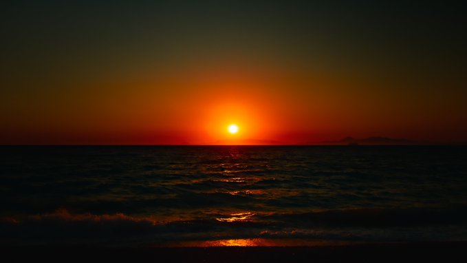 A serene sunset over the ocean, with the bright orange sun low on the horizon casting a glowing reflection across the dark, rippling water.