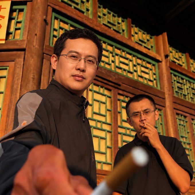 Two men stand in front of a traditional Chinese wooden lattice facade, with the man in the foreground wearing glasses and a dark jacket while the second man in the background smokes a cigarette, fitting the post's reference to "Wanna have a Hua Zi?" (a popular Chinese cigarette brand). The setting appears to be an evening scene with warm lighting against the ornate architectural background.