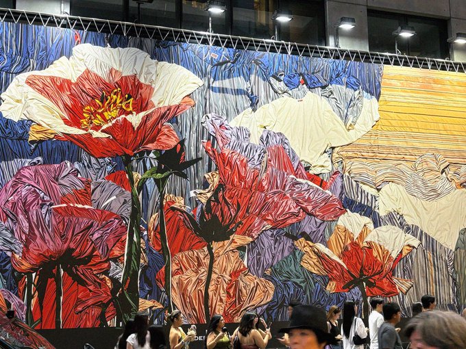A large, colorful textile or mural depicting oversized flowers, including a prominent red poppy, displayed as people walk in front of it.