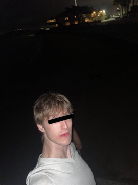 A nighttime selfie of a young man with blonde hair wearing a light-colored hoodie, with his eyes censored by a black bar, standing in a dark outdoor setting with distant lights.