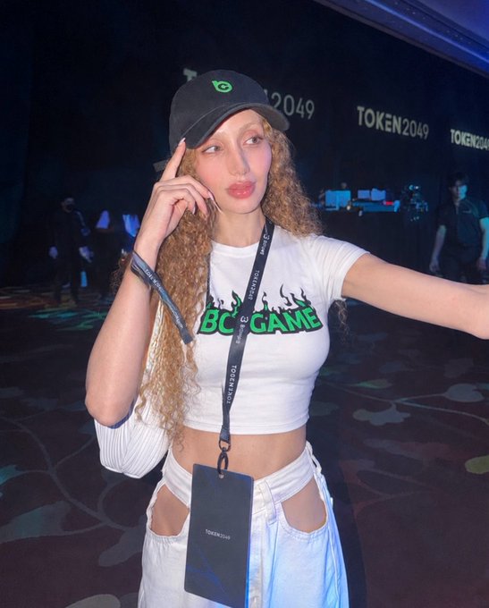 A young woman with curly blonde hair wearing a black cap and a white 'BC GAME' crop top poses at the TOKEN2049 crypto conference, with a lanyard and event badge around her neck.