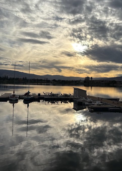 A serene sunset scene over a lake with boats docked at a pier, the sky and clouds beautifully reflected in the calm water.