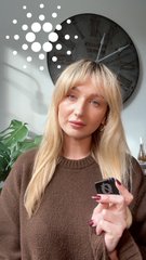 A blonde woman indoors faces the camera while holding a small black device, with a wall clock and decorative lights behind her.