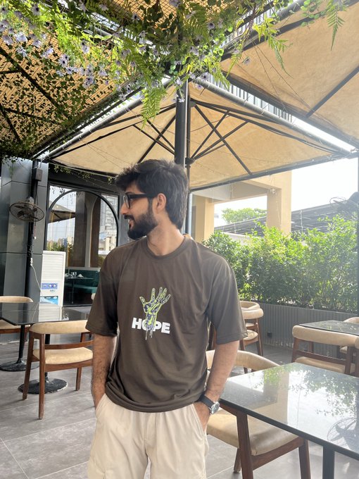 A bearded man with glasses stands under a canopy of hanging vines, wearing a brown t-shirt featuring a skeleton hand graphic and the word "HOPE."