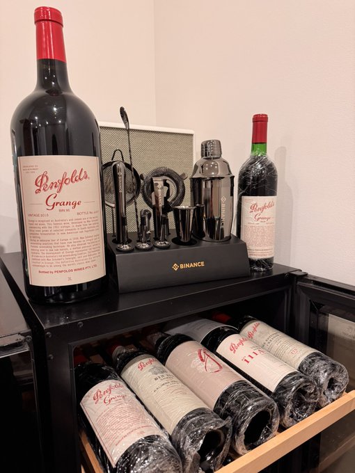 A bottle of Penfolds Grange Shiraz 2017 next to a cocktail shaker and bar tools branded with the Binance logo, with additional Penfolds Grange bottles stored horizontally in a wine rack below.