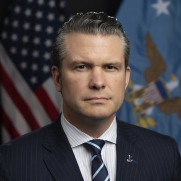 A formal portrait shows a man in a dark pinstripe suit with a striped tie, standing before the American flag and the U.S. Department of Defense flag, identified as Defense Secretary Pete Hegseth in connection with a news story about his disclosed Bitcoin holdings of up to $65,000 in a Coinbase wallet.