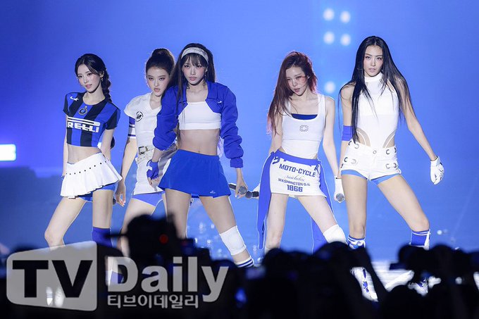 Another stage performance shot of the same K-pop girl group in blue and white racing-inspired outfits, this time with a 'TV Daily' watermark.