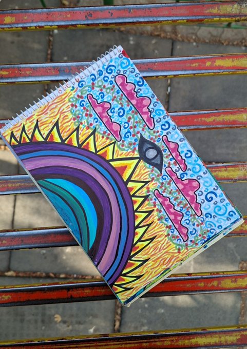 A spiral notebook featuring colorful hand-drawn artwork of a rainbow, sun, and abstract patterns, resting on a red and blue painted metal bench.