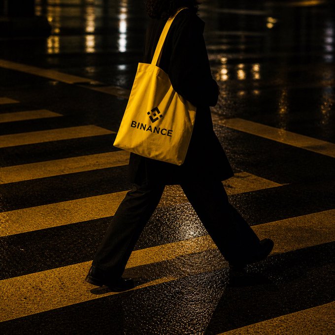 A nighttime photo of a person walking across a zebra crossing carrying a yellow tote bag with the "BINANCE" logo.