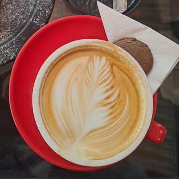 A top-down view of a latte with intricate leaf-shaped foam art, served in a red cup and saucer alongside a chocolate cookie.