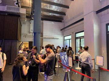 A line of people waiting at what appears to be a store or event entrance, separated by a stanchion rope, in a modern industrial-style interior.