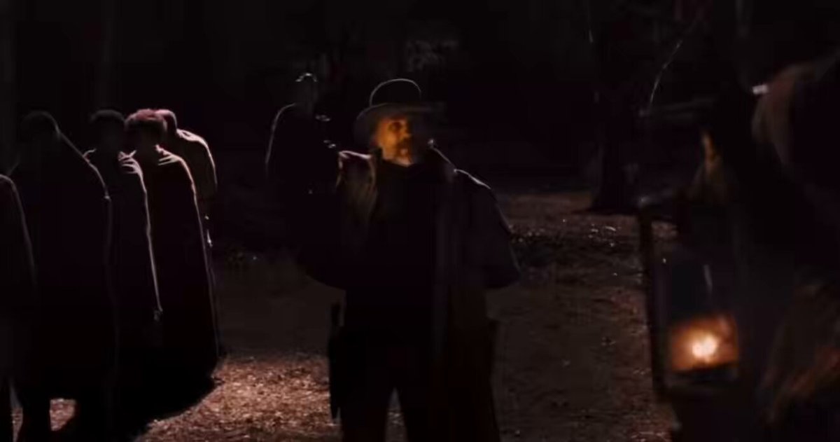 The image shows a group of people standing around, with one person holding a lantern. The scene appears to be set in a dark forest or alleyway at night. There are several individuals present, including a man wearing a hat who seems to be the main focus of the photo. The other people in the picture appear to be either walking by or standing still, creating an atmosphere of mystery and intrigue.