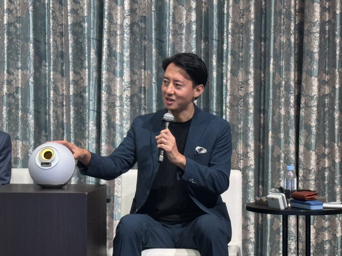 A man in a navy blazer speaks into a microphone while gesturing toward a white spherical device (likely the Orb used for World ID verification), seated on stage at the Nikkei-hosted "GEN AI Summit" panel discussion.