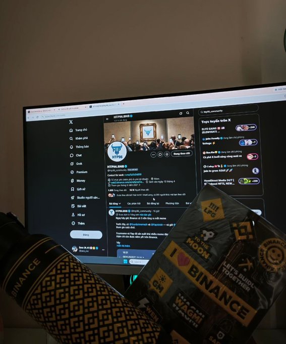 A computer monitor displaying the X/Twitter profile of @HTP98_bnb alongside Binance-branded merchandise and promotional items on a desk.