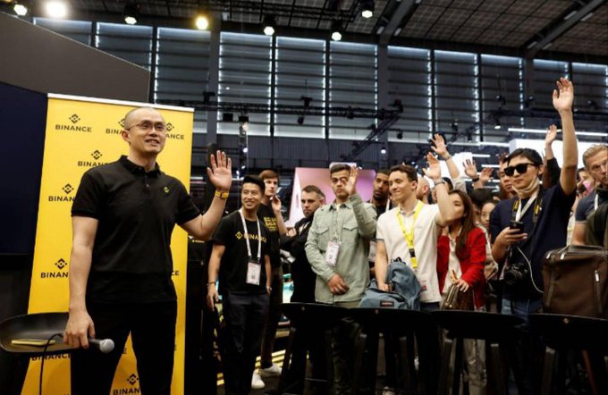 Changpeng "CZ" Zhao waves to an enthusiastic crowd at a Binance-branded event, with the company's yellow logo backdrop visible behind him. The image illustrates his active public role in promoting crypto industry initiatives, including his support for an ASEAN "passport" system that would let licensed crypto firms expand more easily across Southeast Asian markets.