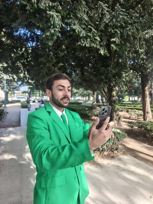 A young man with a beard wearing a bright green suit and tie, standing on a park pathway and taking a mirror selfie with his smartphone while surrounded by trees.