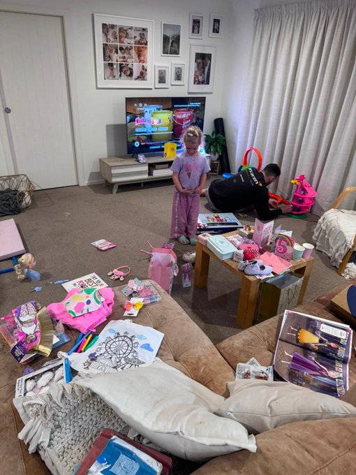 A young girl stands in a living room strewn with the aftermath of a birthday party, surrounded by opened gifts, toys, wrapping paper, and a "Happy Birthday" banner, while a man crouches nearby sorting through presents. The TV in the background displays a Minecraft menu screen, and a toy box labeled with "20+ pieces" sits on the couch. The scene reflects the post-celebration chaos described, marking the birthday of "Lil Shaqita" whose daughter inspired her parent's journey into web3.