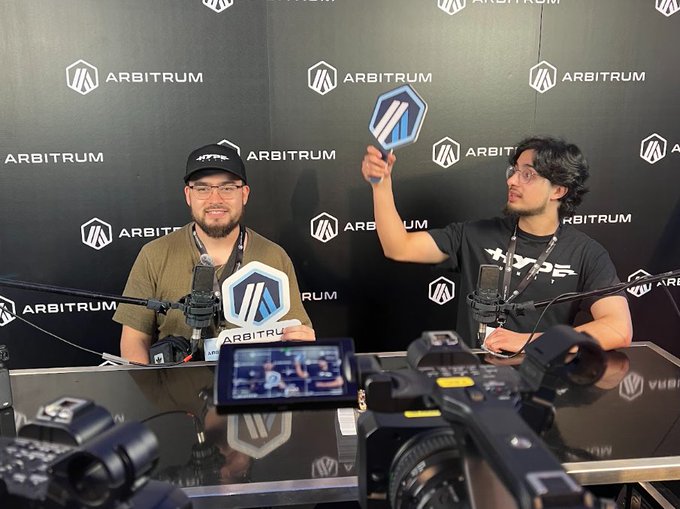 Two men sitting at a podcast or interview setup with 'ARBITRUM' branding in the background, holding Arbitrum-branded signs while cameras record them.