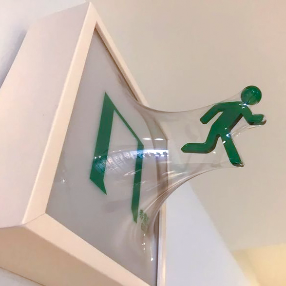 A white emergency exit sign featuring a green running figure and door symbols, with the plastic cover appearing melted or distorted.