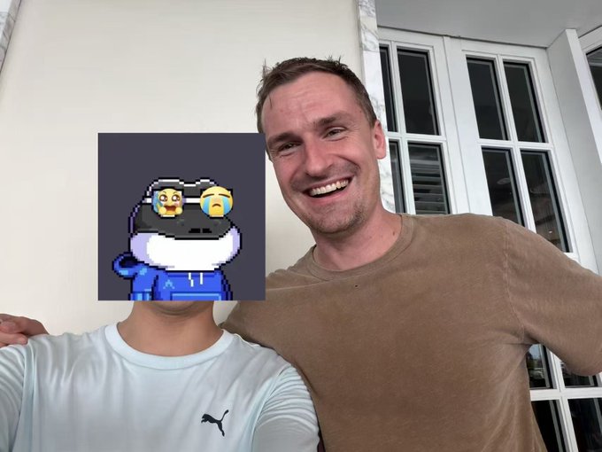 Two men pose for a photo indoors; the man on the left has his face obscured by an anime avatar of a cat wearing a spacesuit.