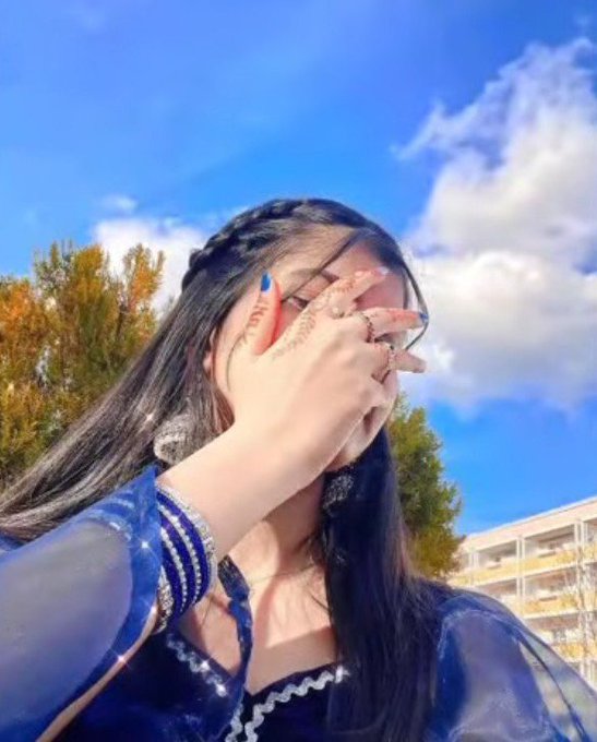A young woman in a blue outfit covers her face with her hand, which has blue nail polish, against a backdrop of blue sky and autumn trees.