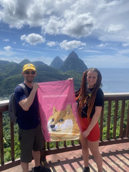 A man in a yellow cap and a woman with dreadlocks holding a pink flag featuring a Doge meme on a scenic overlook with Pitons mountains in the background.