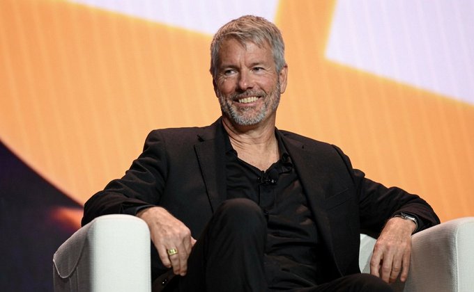 Michael Saylor, the executive chairman of Strategy (formerly MicroStrategy), smiles while seated in a light-colored armchair during what appears to be a speaking engagement or interview, connected to news that his company has increased its USD reserves by $450 million, bringing their total holdings to 843,775 BTC and $3 billion in cash.