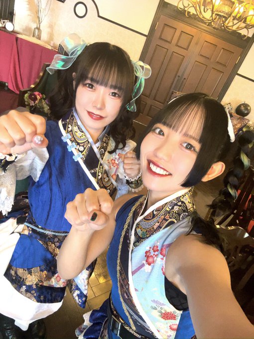 A selfie of two smiling young women with dark hair wearing ornate blue and floral traditional-style outfits, posing with their fists forward in an indoor setting.
