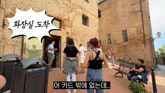 A group of people waiting in line outside a stone building, with a woman with purple hair in a black tank top standing in the foreground; a Korean speech bubble reads "화장실 도착" (Toilet arrival) and subtitles state "어 카드 밖에 없는데.." (Oh, I only have a card..), illustrating the irony of being told to pay with coins while the people in line have only their credit cards.