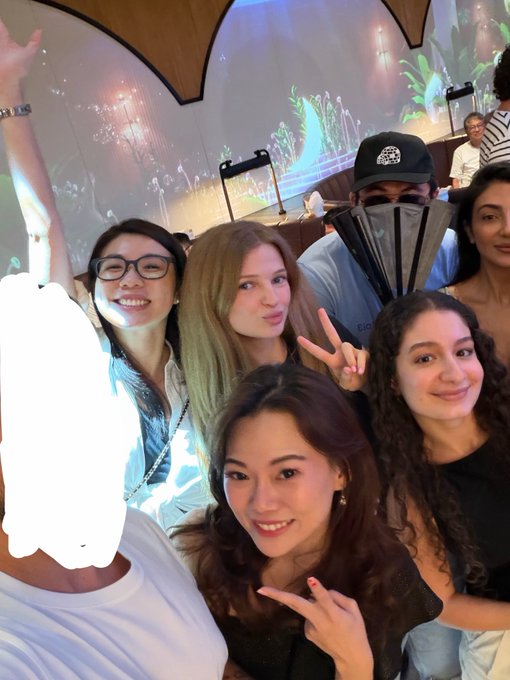 A group selfie of several young women and a man wearing a black cap, posing in what appears to be a venue with decorative lighting in the background.