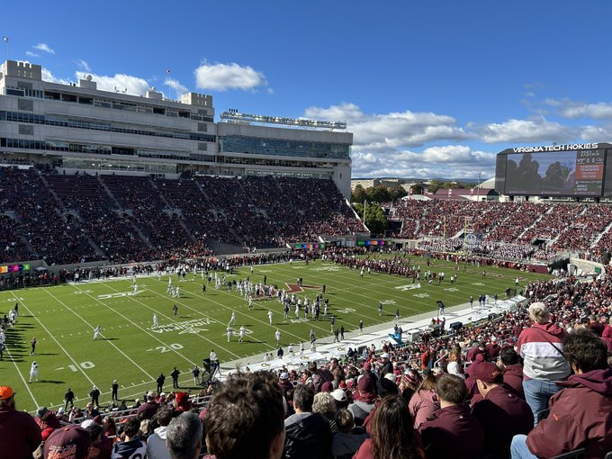 A crowded college football stadium scene with players on the field and a scoreboard reading 'VIRGINIA TECH HOKIES' visible in the upper right.