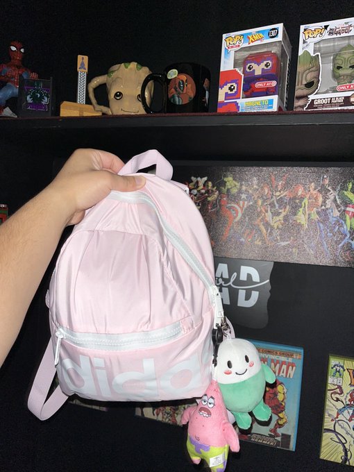 A hand holds up a pink backpack in front of a black shelf displaying Funko Pop figures, mugs, and Marvel comic artwork, with a Patrick Star plush keychain attached.