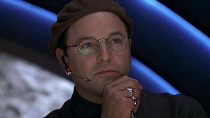 A man wearing round glasses, a tan beret, and a headset microphone rests his chin on his ring-adorned hand with a thoughtful, knowing expression, set against a curved blue-lit backdrop. The image pairs with the post's satirical "strategic announcement" timeline to humorously portray the mastermind scheming behind manipulative crypto project launches.