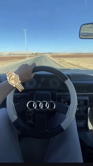 A first-person view from the driver's seat of an Audi, showing a hand wearing a gold Rolex watch on the steering wheel, looking out onto a long, straight rural road under a clear blue sky.