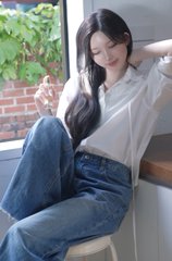A young woman with long dark hair sits by a window in a white shirt and jeans, adjusting her hair while looking downward.