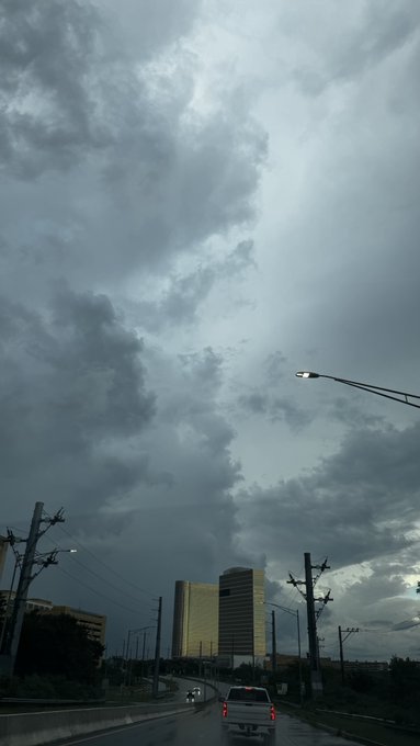A view from inside a car driving on a highway under a dramatic, cloudy sky with sunlight breaking through, passing city buildings and power lines.