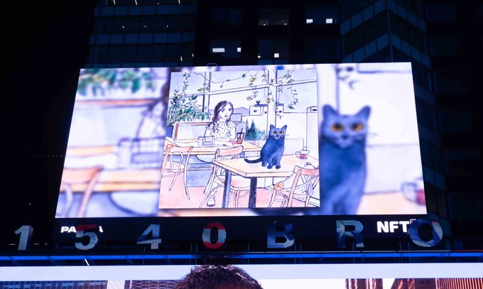 A large outdoor digital billboard displays an animated illustration of a woman sitting at an outdoor café table alongside a black cat, with the text '1540BRO NFT' visible at the bottom.
