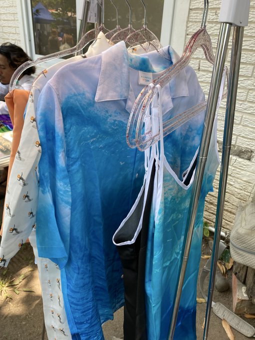 Blue and white tie-dye button-up shirts hang on a clothing rack, with white shirts featuring small animal patterns visible in the background.