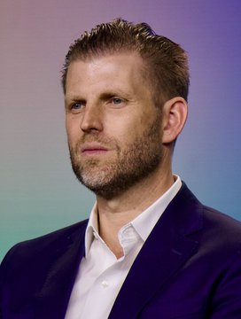 A man with short blondish hair and a light beard, wearing a dark suit jacket over a white collared shirt, is pictured against a softly lit purple-blue backdrop. The image accompanies news of Eric Trump's Bitcoin mining venture, American Bitcoin Corp., wiping out roughly $600 million in family wealth during a downturn in the sector.