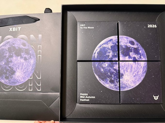 An open black gift box labeled 'XBIT To The Moon 2026' containing four square cards that form a purple moon image, accompanied by text reading 'Happy Mid-Autumn Festival' and a small horned logo.